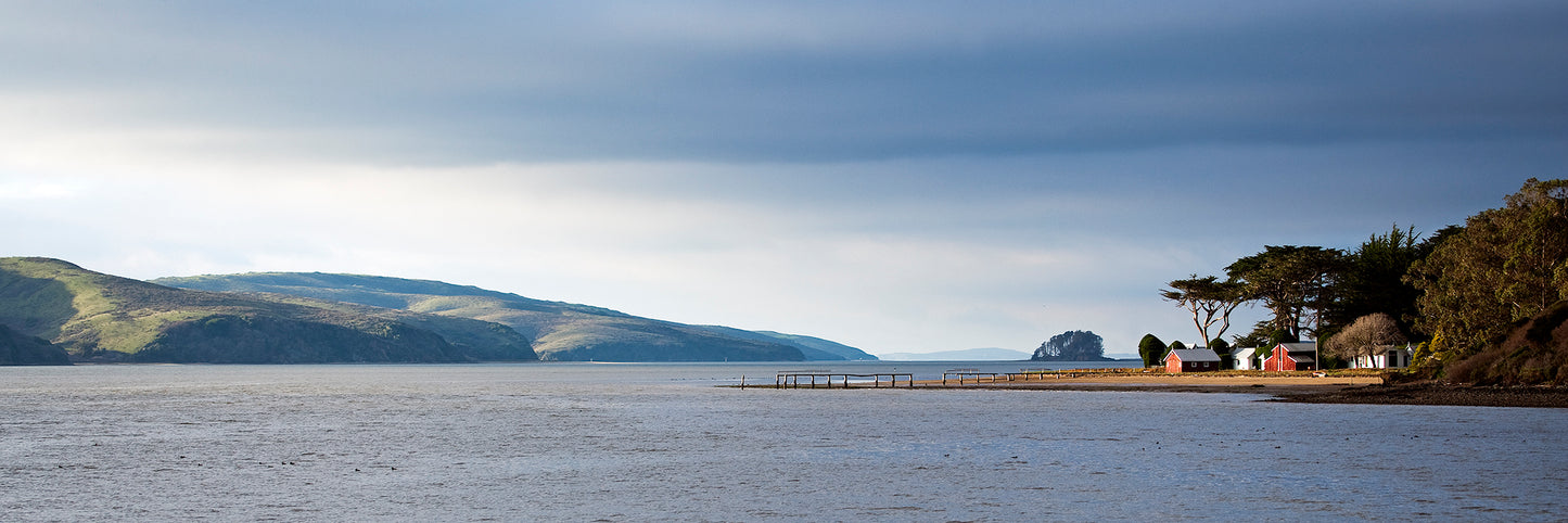 Colorful coastal buildings and cypress trees line the shore of Tomales Bay under dramatic winter skies, with rolling hills and calm water in the distance.