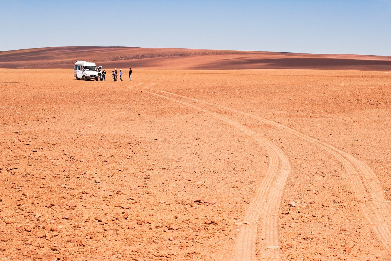 A group of people stands beside a white van under a vast blue sky in the Atacama Desert, with tire tracks curving across the red, rocky terrain toward distant rolling hills.