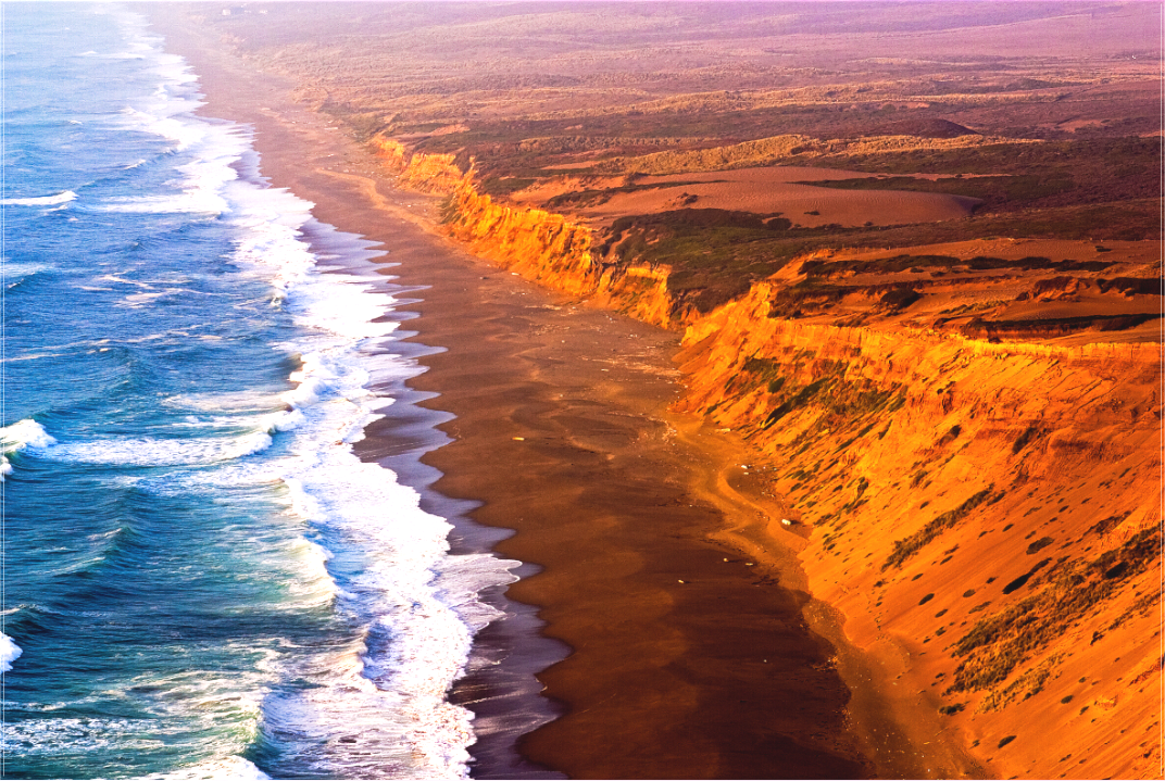 Dramatic Labor Day sunset along the Point Reyes Coastline, with golden light illuminating rugged sea cliffs and surf crashing onto the remote beach below, viewed from a high cliff near the historic lighthouse.