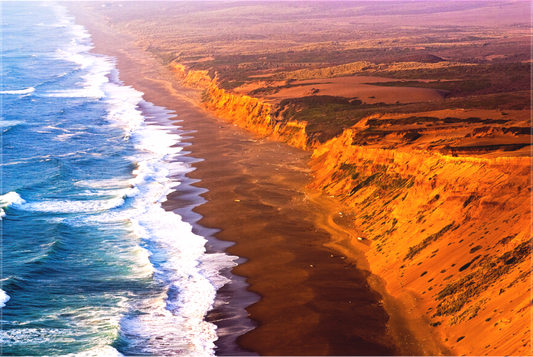 Dramatic Labor Day sunset along the Point Reyes Coastline, with golden light illuminating rugged sea cliffs and surf crashing onto the remote beach below, viewed from a high cliff near the historic lighthouse.