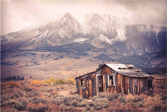 A weathered, abandoned wooden cabin stands in the foreground of an autumn landscape east of Tioga Pass near Mono Lake, with dramatic snow-dusted peaks rising behind under misty skies; texture and vignetting enhance the vintage, moody atmosphere.