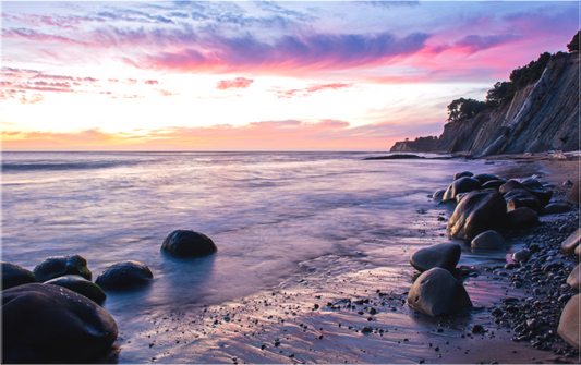 Smooth, round boulders scatter across the shore at Bowling Ball Beach during a vibrant fall sunset, as gentle waves wash over the sand and pastel clouds streak across the sky at Schooner Gulch.
