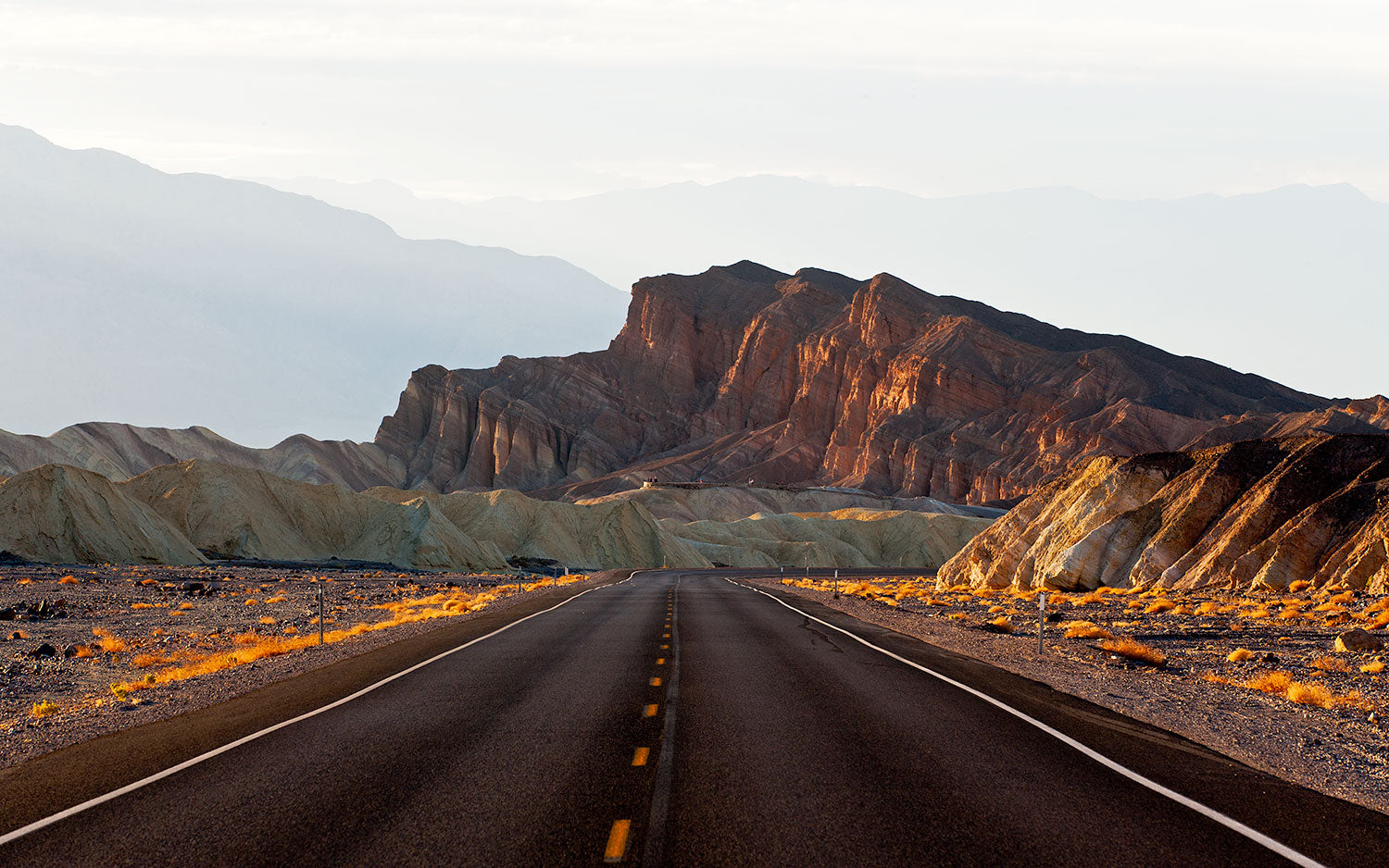 A straight road leading through a desert landscape toward the golden-hued badlands and rugged formations near Zabriskie Point, captured on a spring afternoon in Death Valley.