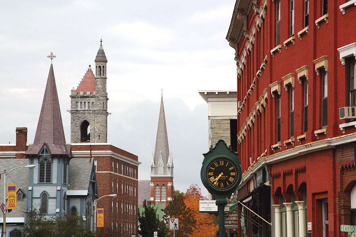 A picturesque street scene in St. Johnsbury, Vermont, captured on a fall afternoon during peak foliage, featuring historic brick buildings, church spires, and a vintage street clock under overcast skies.