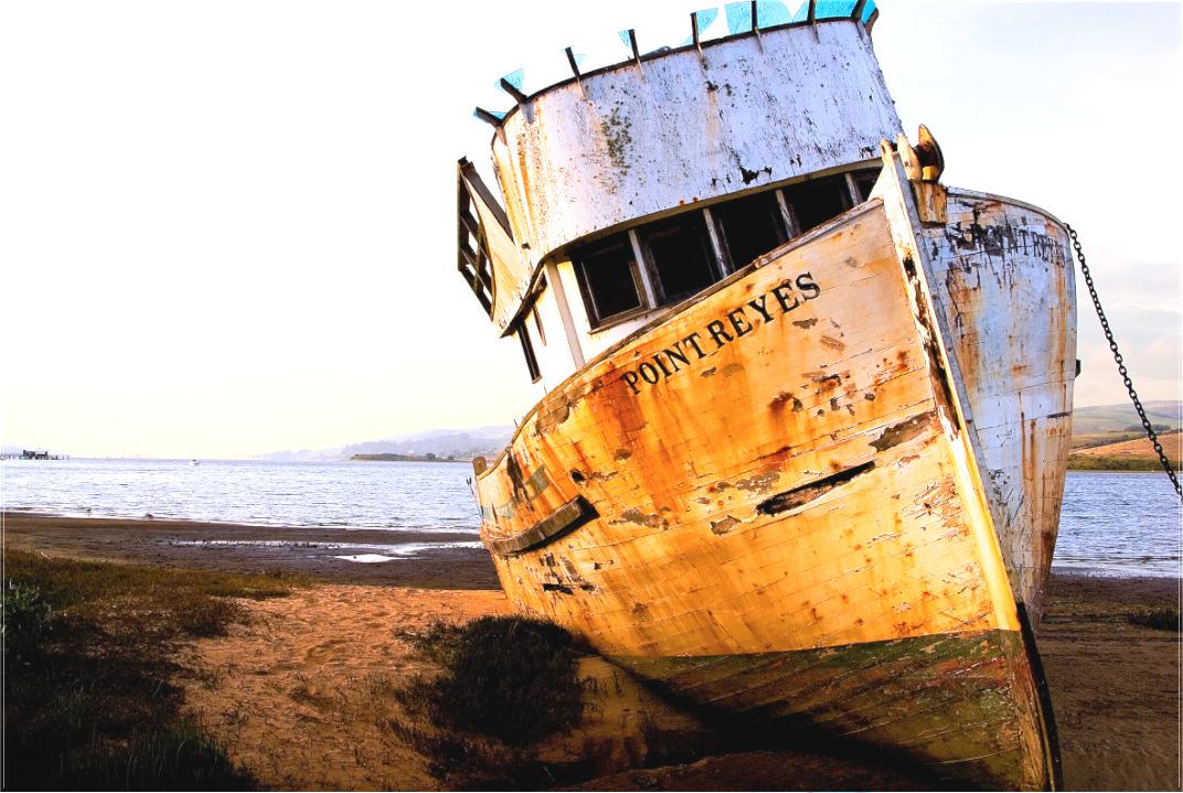 Weathered fishing boat named “Point Reyes” leaning on the shore of Tomales Bay, photographed over 20 years ago when its hull and cabin were still mostly intact, bathed in warm evening light.
