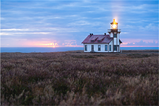 A serene summer sunset at Point Cabrillo Light Station on the Mendocino Coast, with the lighthouse beacon shining warmly as twilight colors reflect over the ocean; once featured as the homepage image for the California State Parks Association.