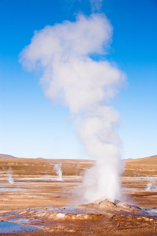 Steam rises from an erupting geyser at El Tatio Geyser Field in Chile, surrounded by a high-altitude desert landscape under a clear blue sky.