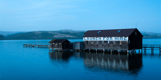 A rustic boathouse with “LAUNCH FOR HIRE” painted across its side sits on stilts over calm blue waters during Blue Hour on Tomales Bay, with rolling coastal hills in the background.