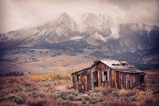 A weathered, abandoned wooden cabin stands in the foreground of an autumn landscape east of Tioga Pass near Mono Lake, with dramatic snow-dusted peaks rising behind under misty skies; texture and vignetting enhance the vintage, moody atmosphere.