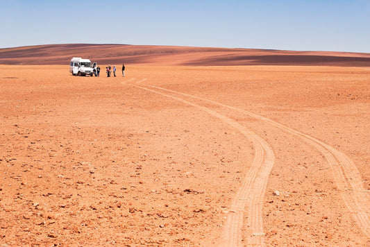 A group of people stands beside a white van under a vast blue sky in the Atacama Desert, with tire tracks curving across the red, rocky terrain toward distant rolling hills.
