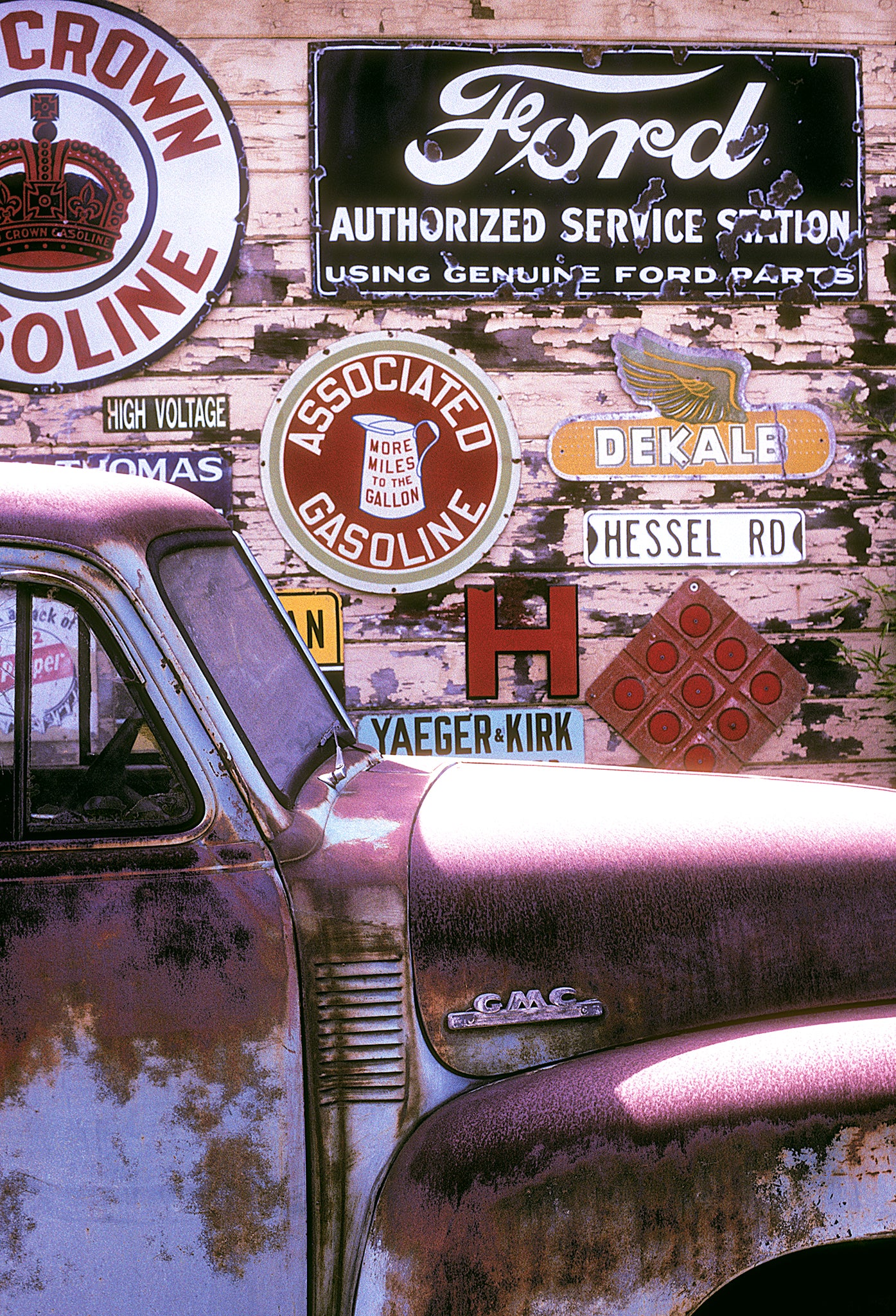 Rusty vintage GMC truck parked in front of a weathered wooden wall covered in old gas station and service signs, photographed in spring on a Sonoma County backroad with Kodachrome film.