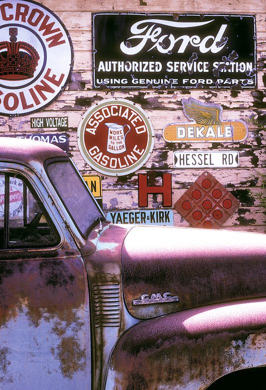 Rusty vintage GMC truck parked in front of a weathered wooden wall covered in old gas station and service signs, photographed in spring on a Sonoma County backroad with Kodachrome film.
