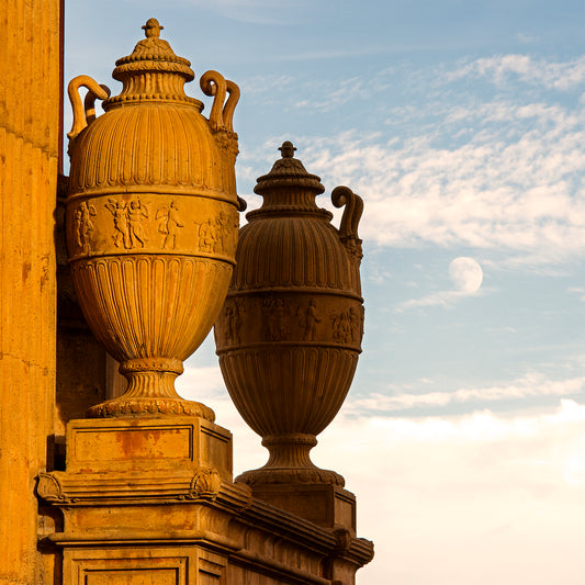Classical urns at the Palace of Fine Arts in San Francisco glow in golden light, their intricate reliefs evoking scenes from antiquity, with the moon rising softly in a pastel sky behind.