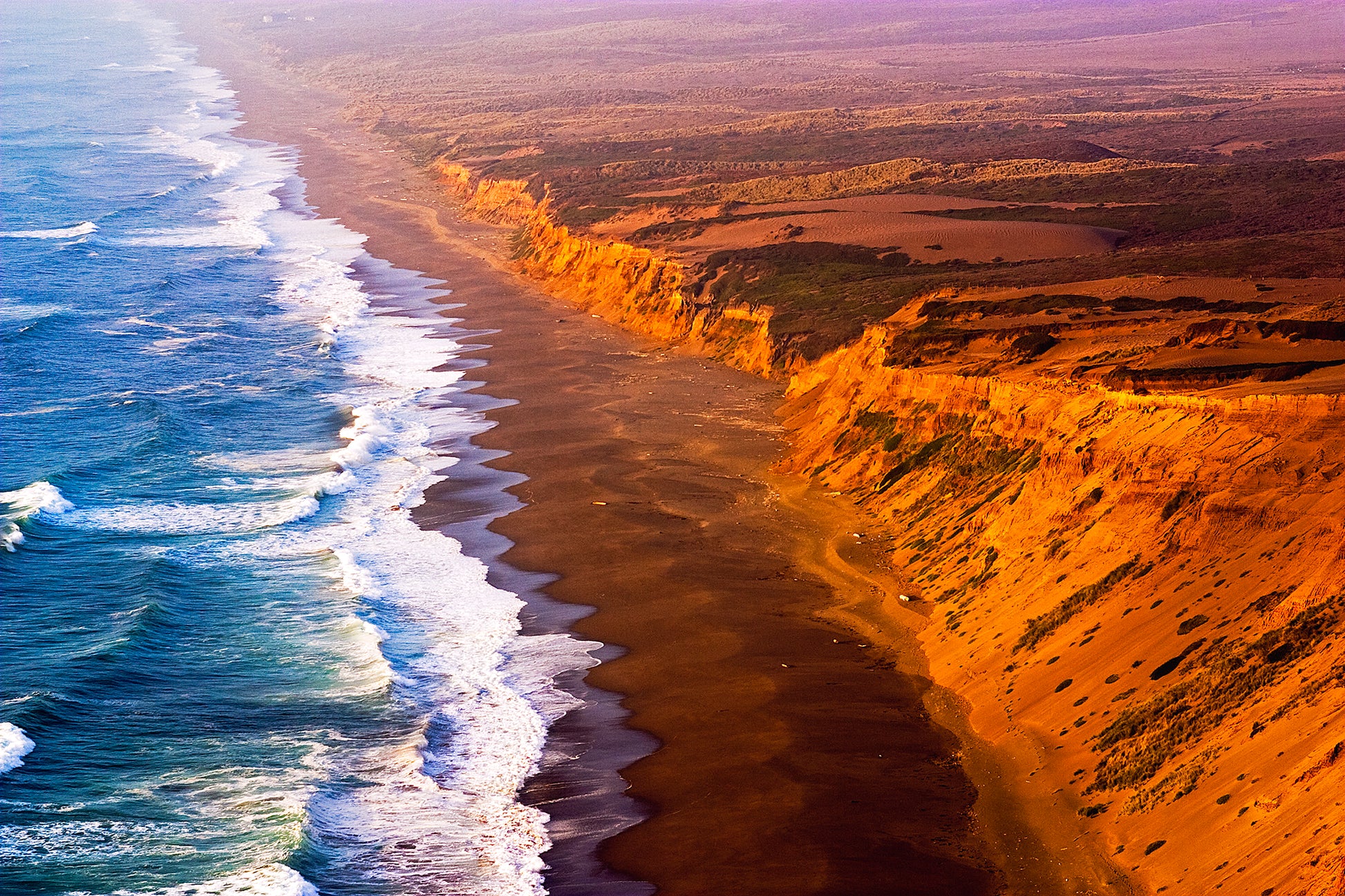 Dramatic Labor Day sunset along the Point Reyes Coastline, with golden light illuminating rugged sea cliffs and surf crashing onto the remote beach below, viewed from a high cliff near the historic lighthouse.