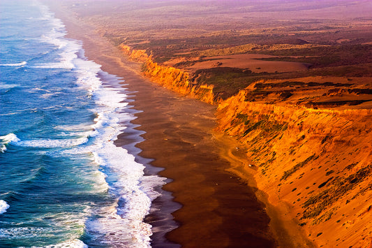 Dramatic Labor Day sunset along the Point Reyes Coastline, with golden light illuminating rugged sea cliffs and surf crashing onto the remote beach below, viewed from a high cliff near the historic lighthouse.