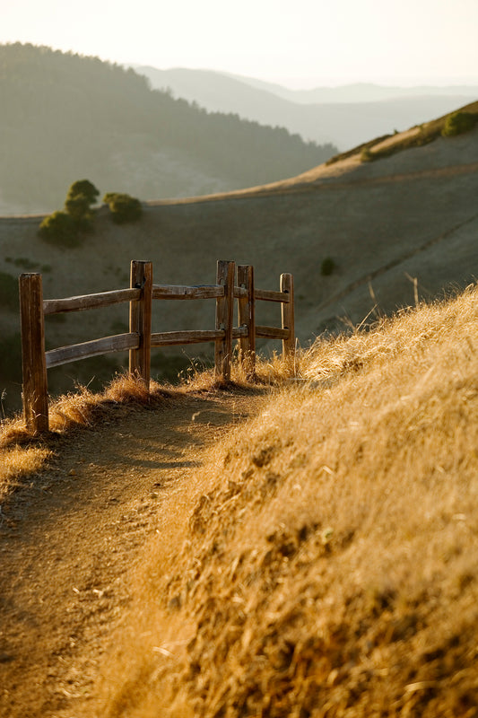Golden light falls on a narrow dirt path and rustic wooden fence along the Big Rock Trail near Skywalker Ranch, with rolling hills and layered ridgelines in the distance on a late Halloween afternoon.