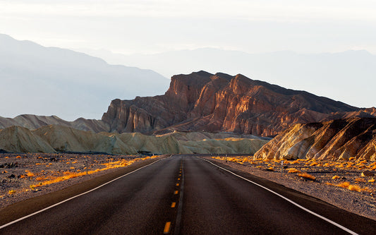 A straight road leading through a desert landscape toward the golden-hued badlands and rugged formations near Zabriskie Point, captured on a spring afternoon in Death Valley.
