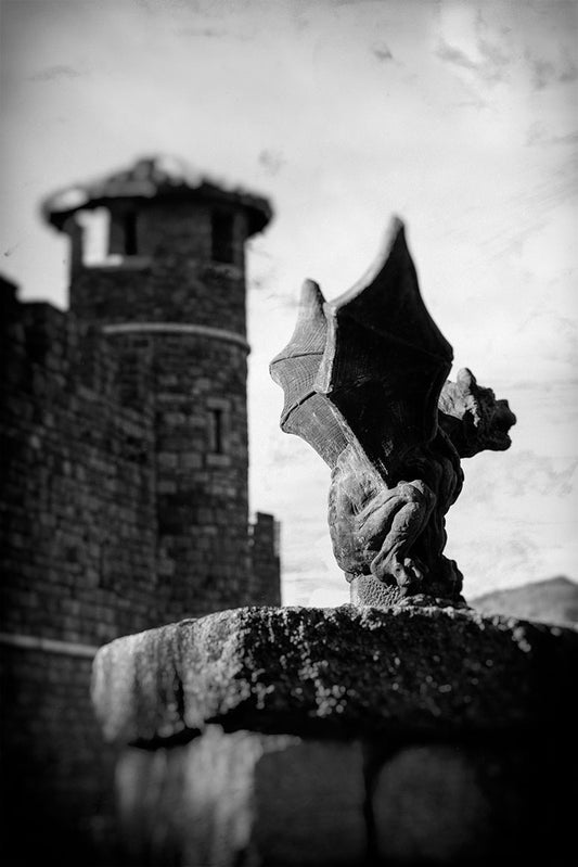 Vintage-style black and white image of a winged gargoyle statue perched on stone, guarding the entrance to a Napa Valley winery designed like a medieval castle.