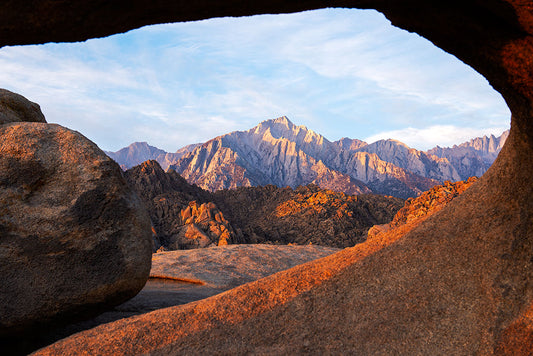 View of the Sierra Nevada mountains at sunrise framed by the Mobius Arch in the Alabama Hills, with warm morning light casting a golden glow on the rugged granite landscape.